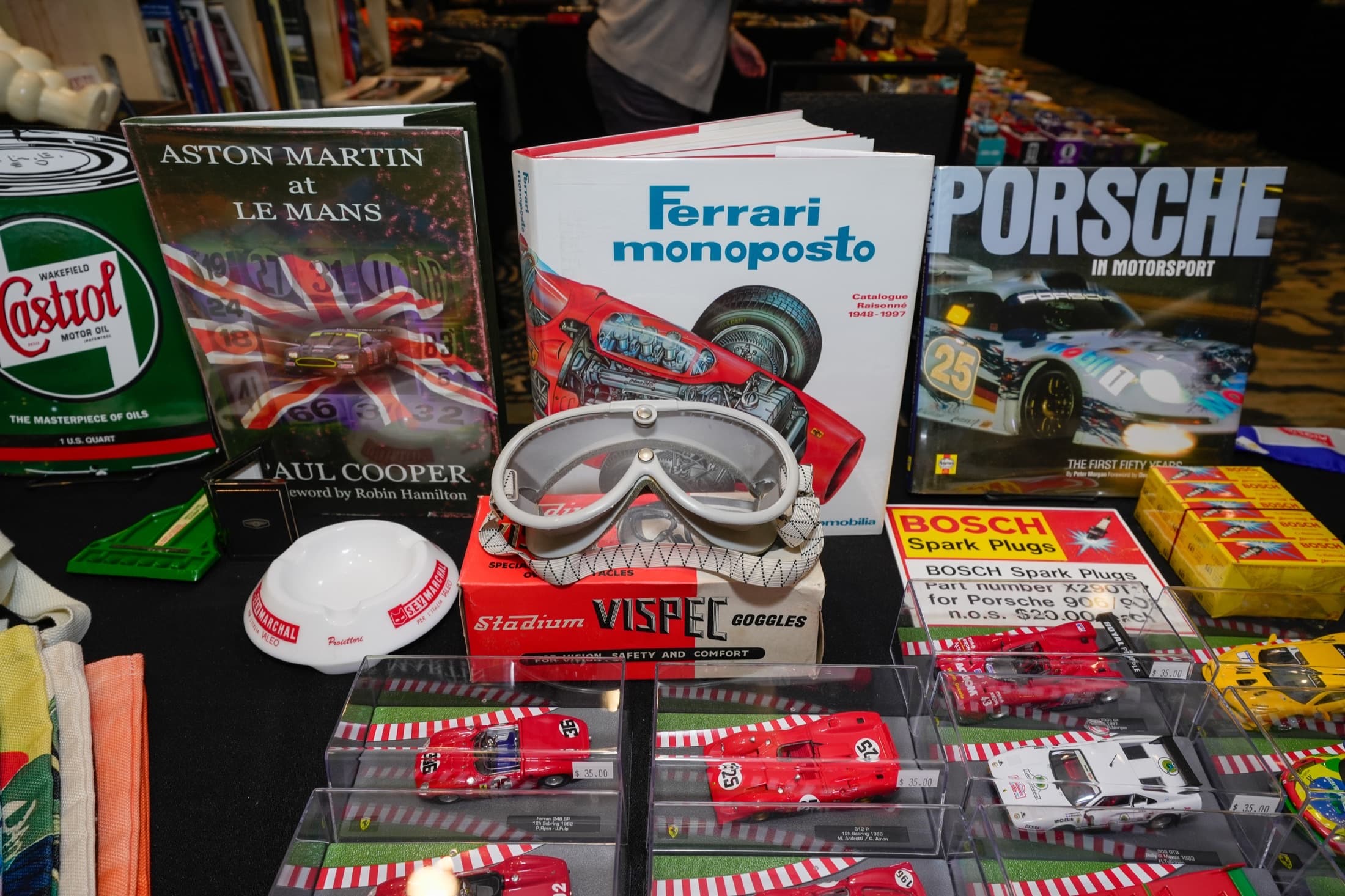 Automobilia display with marque books, scale models, racing goggles, Castrol oil can, and Bosch spark-plug boxes on a black-clothed table at an indoor collector expo