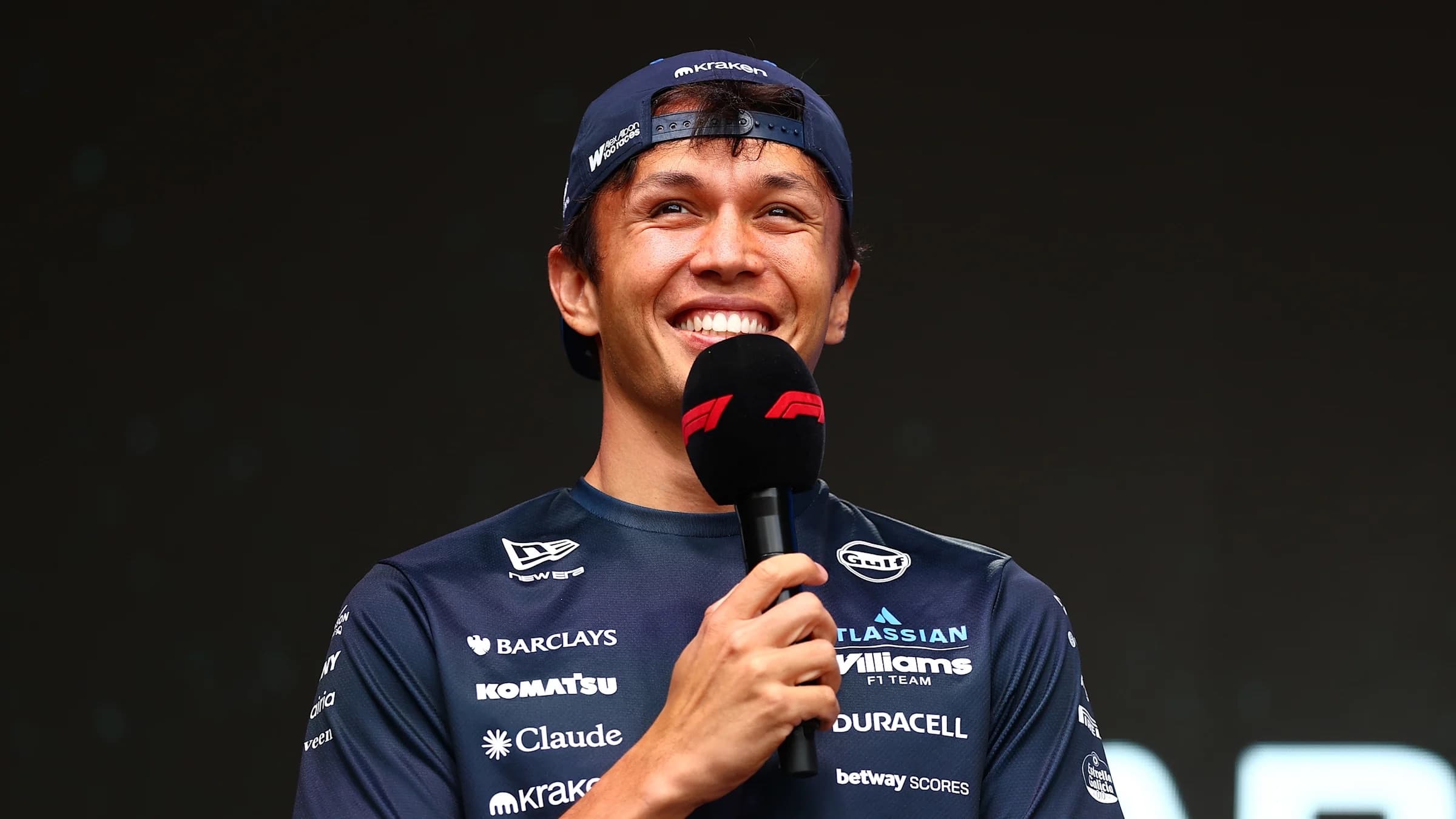Alex Albon smiling in a Williams team shirt and backward cap marked Alex Albon 100 Races while holding an F1 microphone