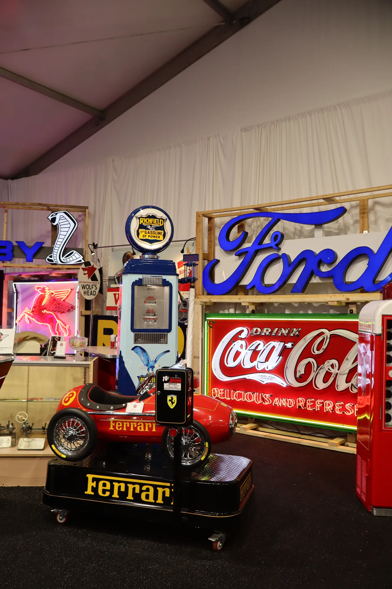 Neon Ford, Coca-Cola, and Shelby signs with a Richfield gas pump and red Ferrari kiddie ride inside the Concours Village memorabilia tent