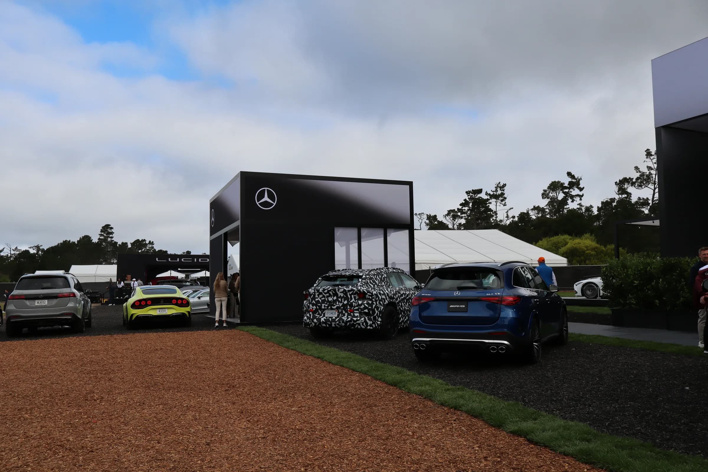 Mercedes-Benz pavilion at Concours Village with an AMG GLC, a camouflaged prototype, and other brand tents under Monterey pines
