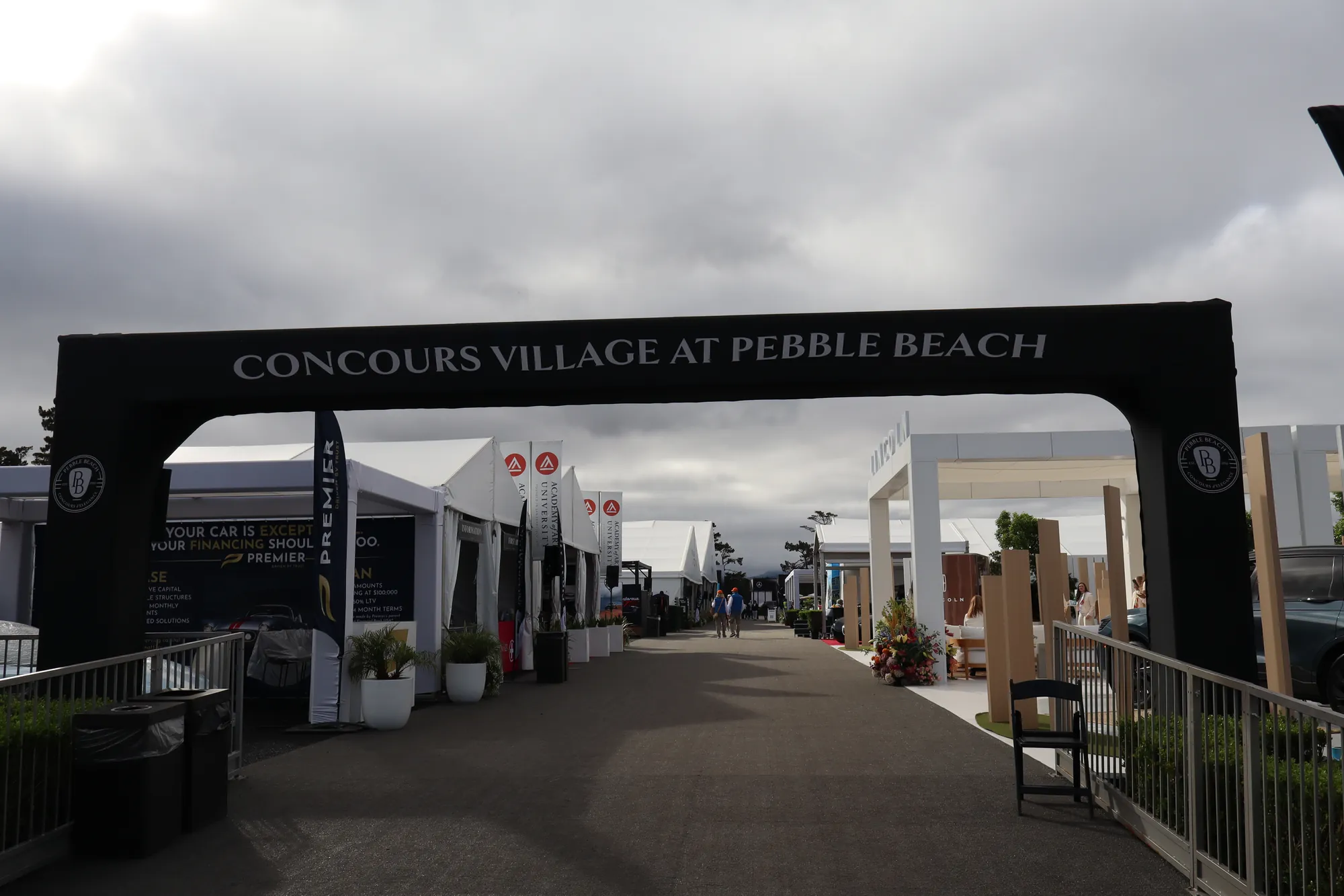 Black Concours Village at Pebble Beach entrance arch looking down a lane of brand pavilions under an overcast sky