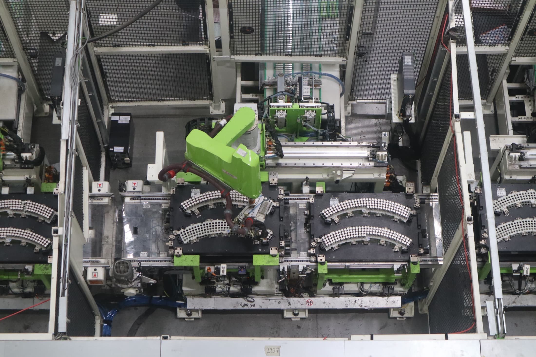 Overhead view of cylindrical battery cells on trays at an electric-scooter battery assembly line with a green robot arm and factory wiring