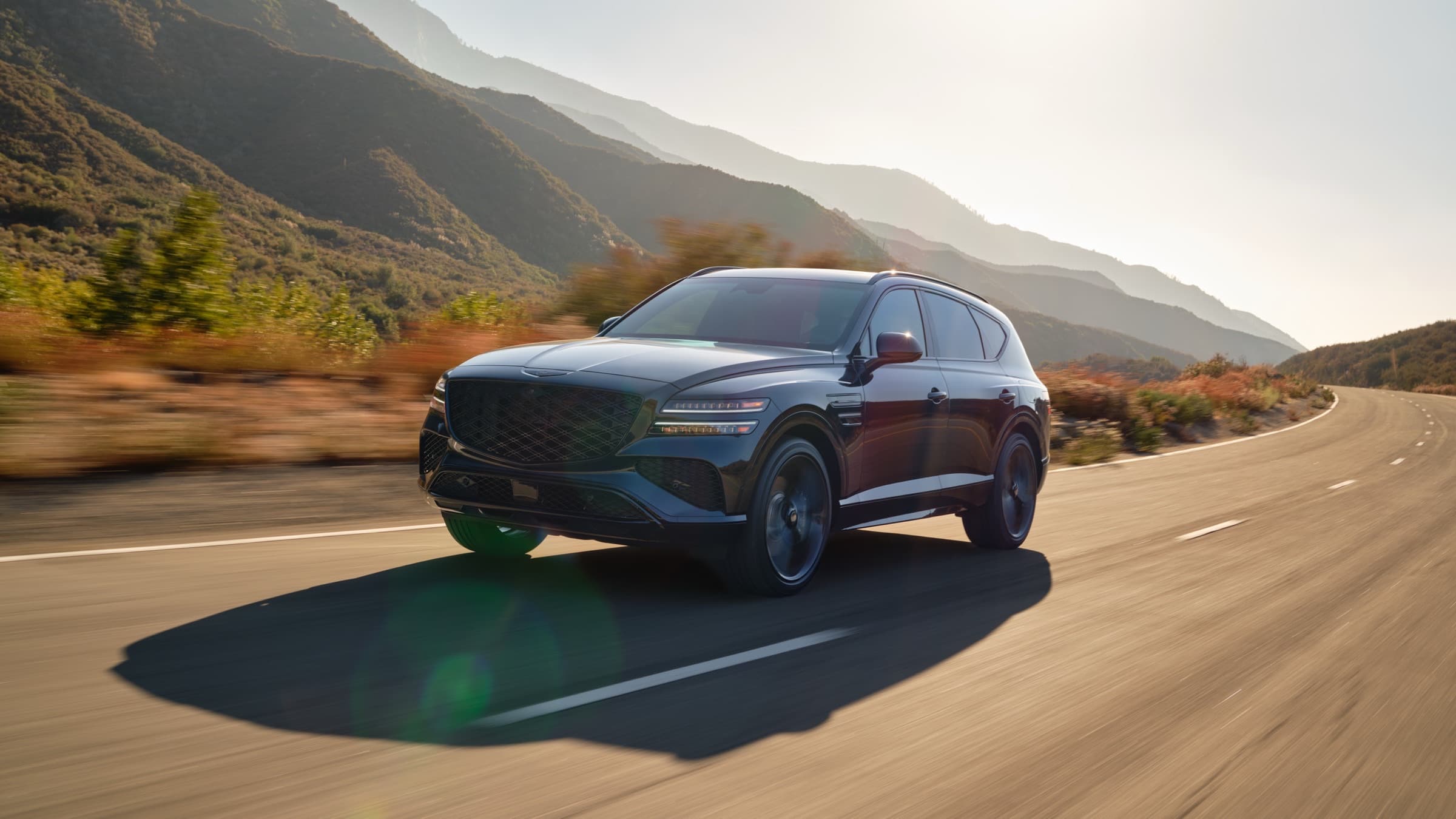 Dark Genesis SUV on a sunlit mountain highway, three-quarter front view in motion, the brand’s crest grille and two-line lamps visible