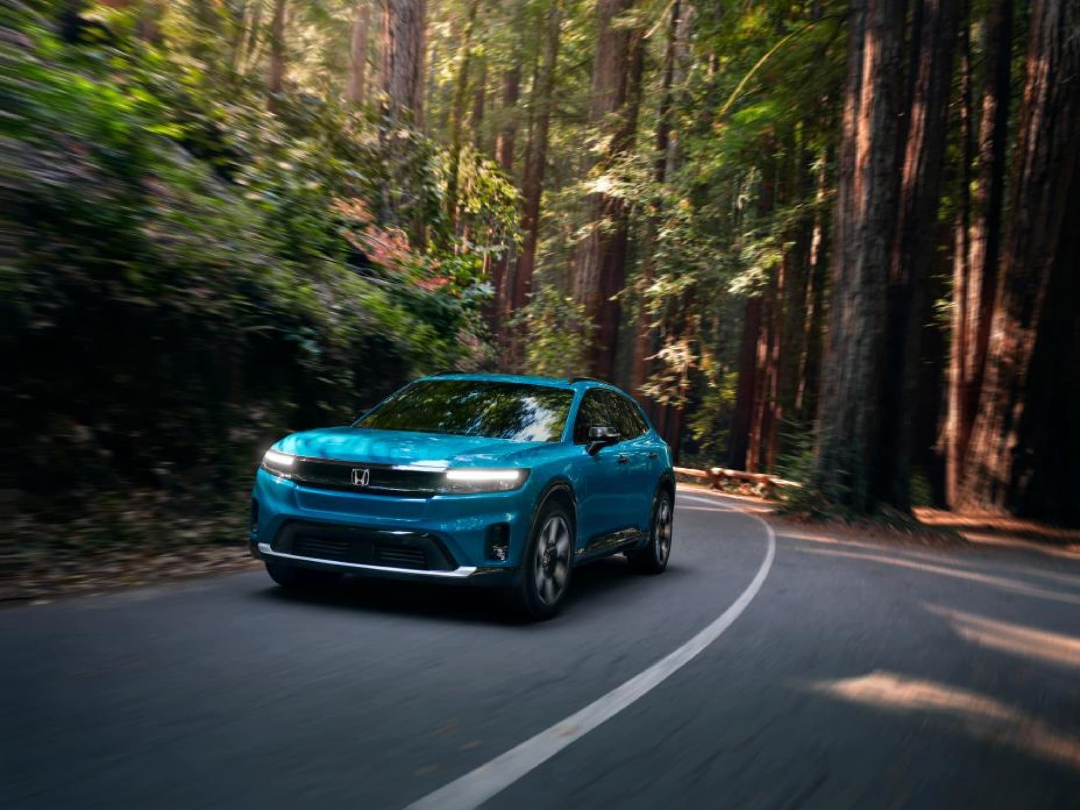 Blue 2025 Honda Prologue Elite driving a forested two-lane road among tall redwoods