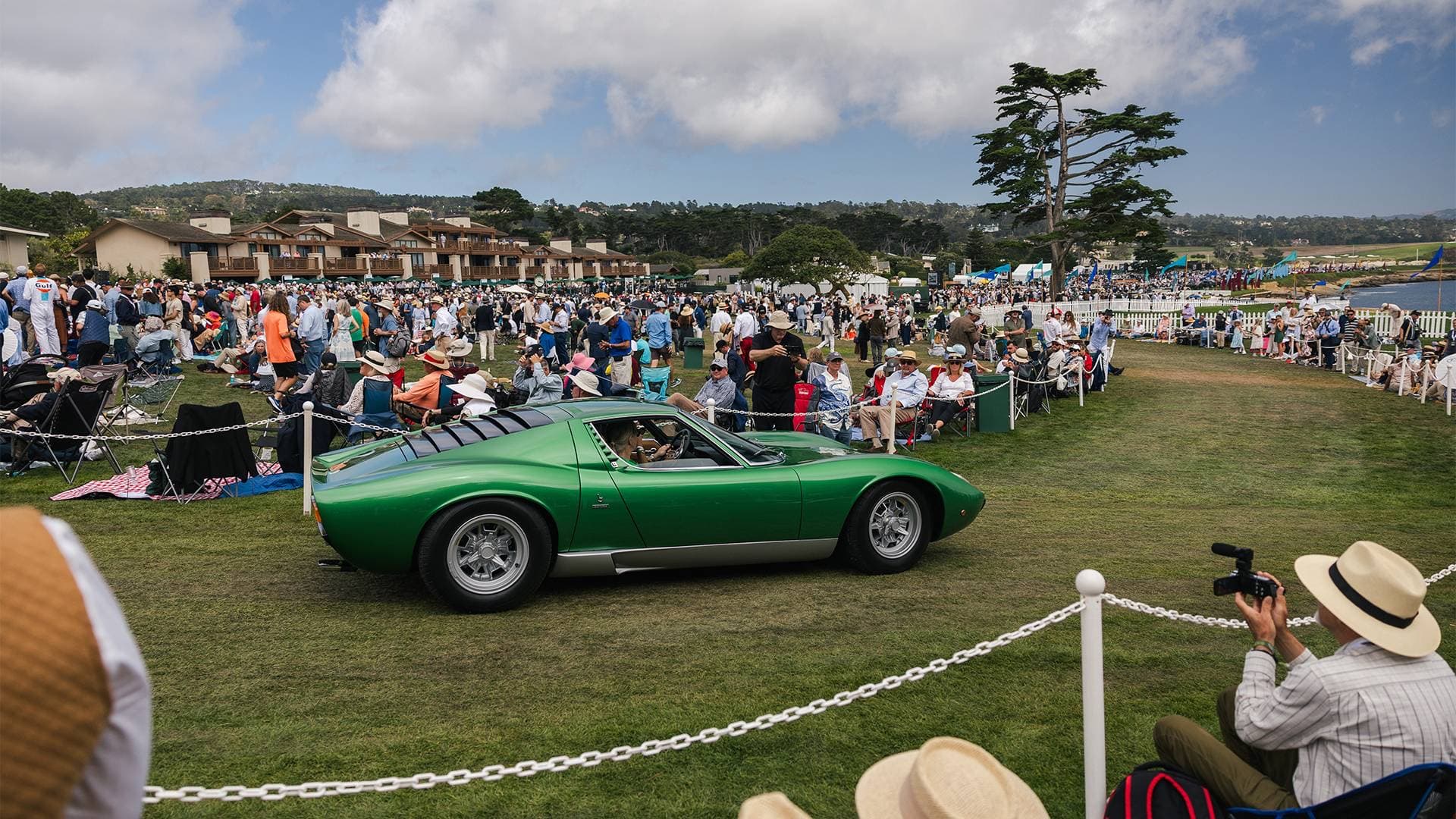 Green Lamborghini Miura driving past spectators on the lawn at Pebble Beach with The Lodge and Monterey Cypress behind