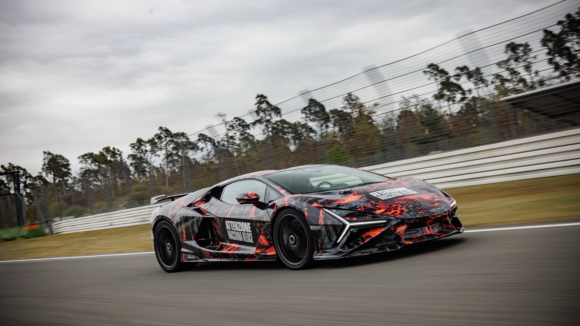 Camouflaged Lamborghini Revuelto SV prototype on track in red-black wrap with ATTENZIONE MACCHINA VELOCE lettering, fixed rear wing, and Y-shaped daytime running lights