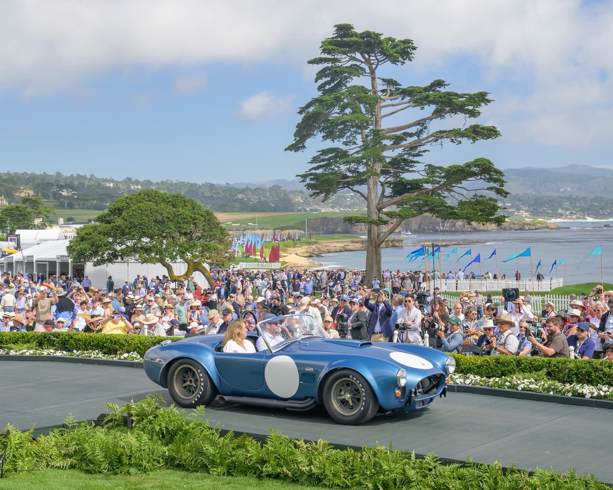 Blue Shelby Cobra on the Pebble Beach Concours awards ramp with spectators, Monterey cypress, and Pacific coastline beyond