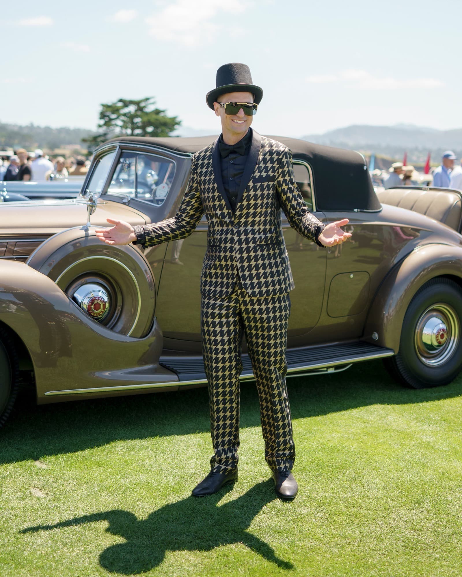 Guest in a gold-and-black houndstooth suit, top hat, and sunglasses posing with arms open beside a Packard Twelve on the Pebble Beach Concours lawn