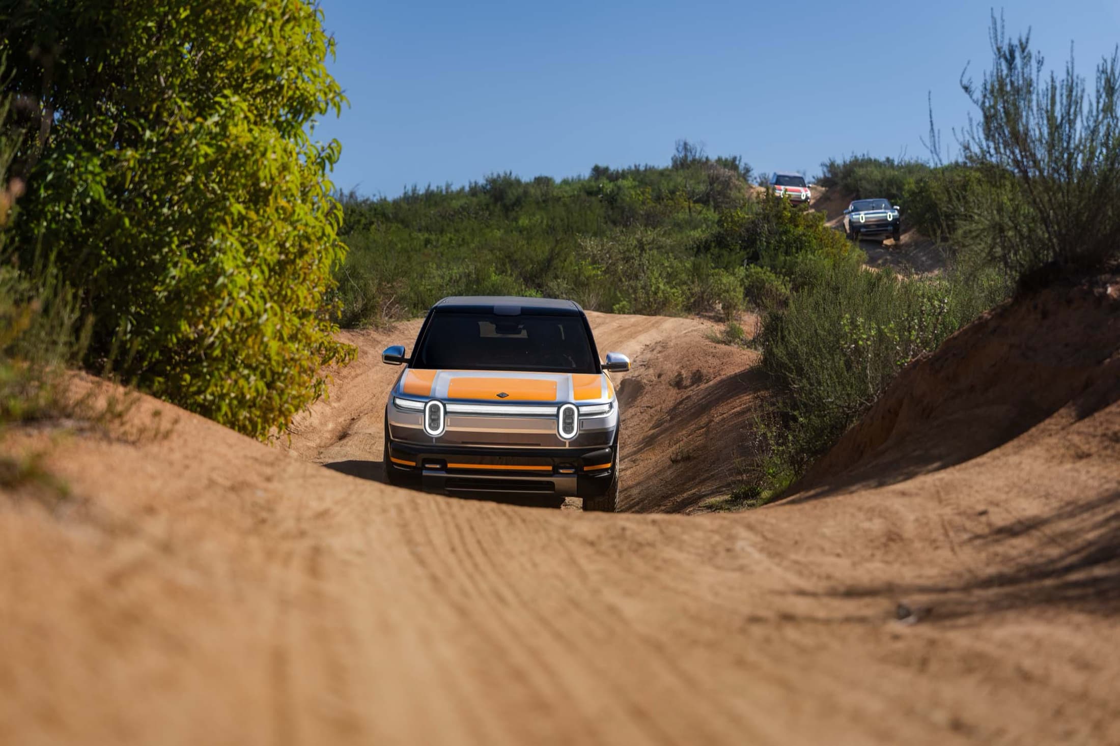Silver and orange Rivian R2 leading a convoy on a sandy off-road trail