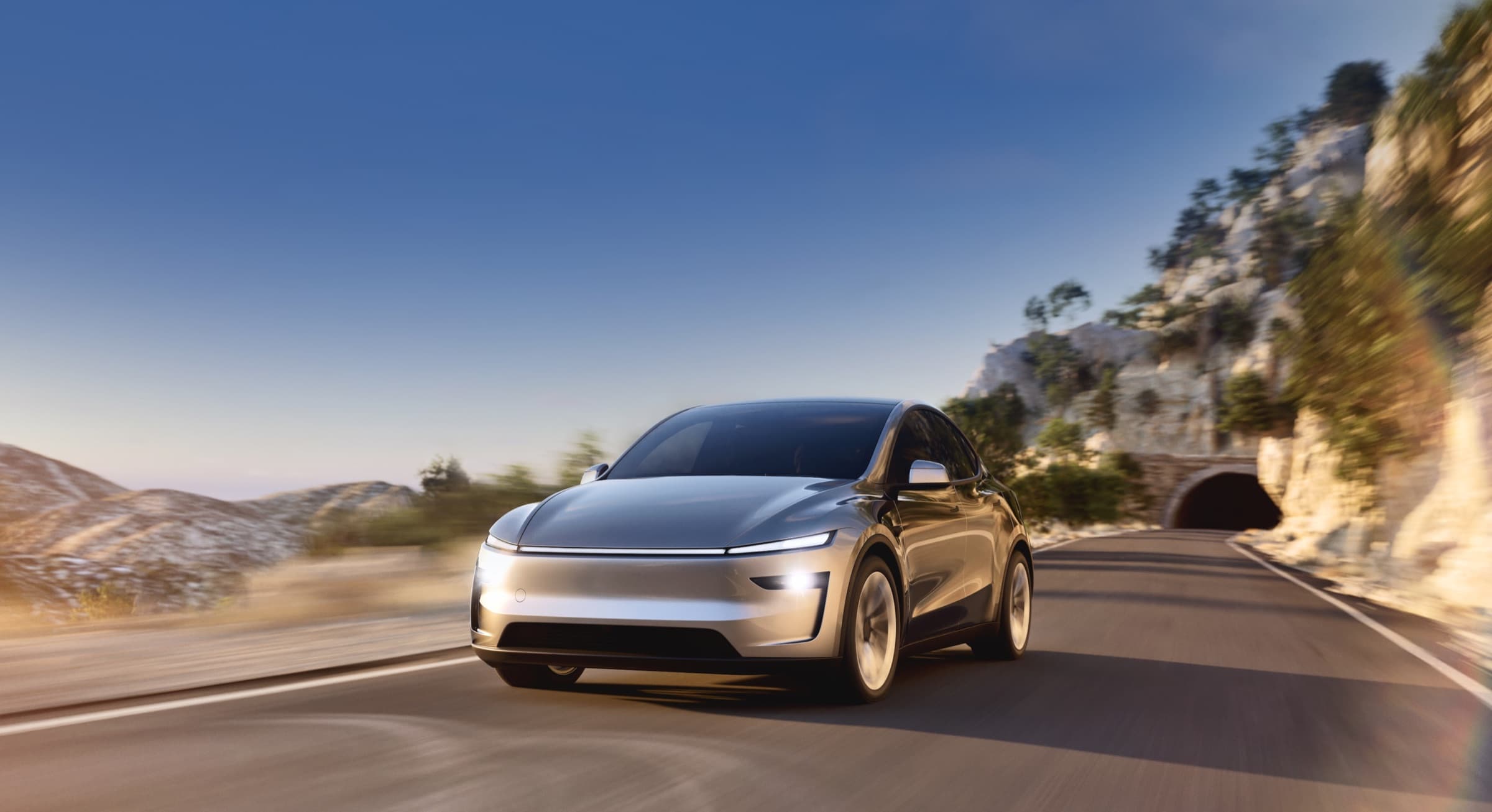 Silver Tesla Model Y driving on a sunlit mountain road near a tunnel entrance