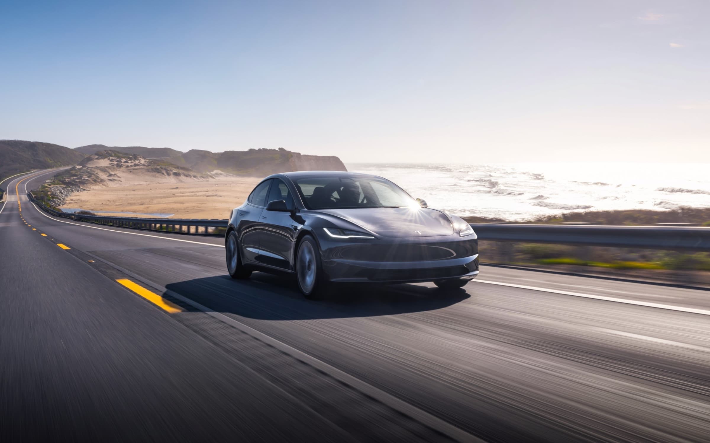 Dark gray Tesla Model 3 driving on a coastal highway, front three-quarter view
